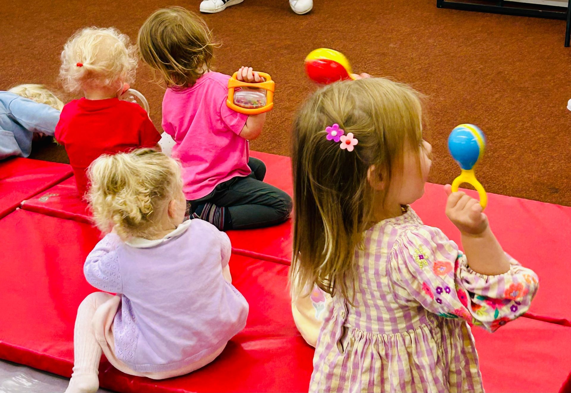 Children sitting on a red mat, playing with colorful handheld musical instruments.