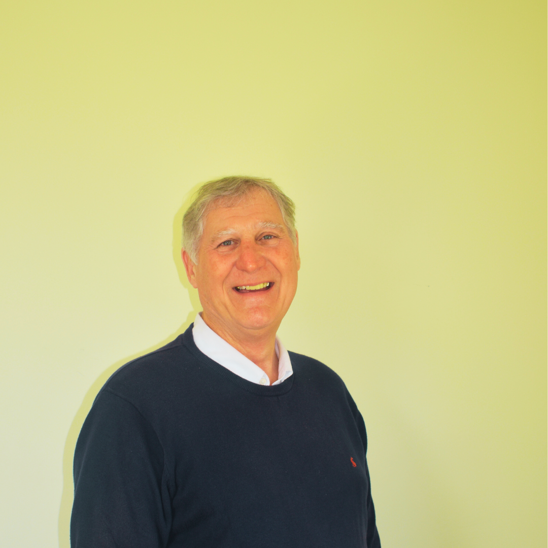 A smiling person wears a navy sweater over a white collared shirt against a plain, light yellow background.
