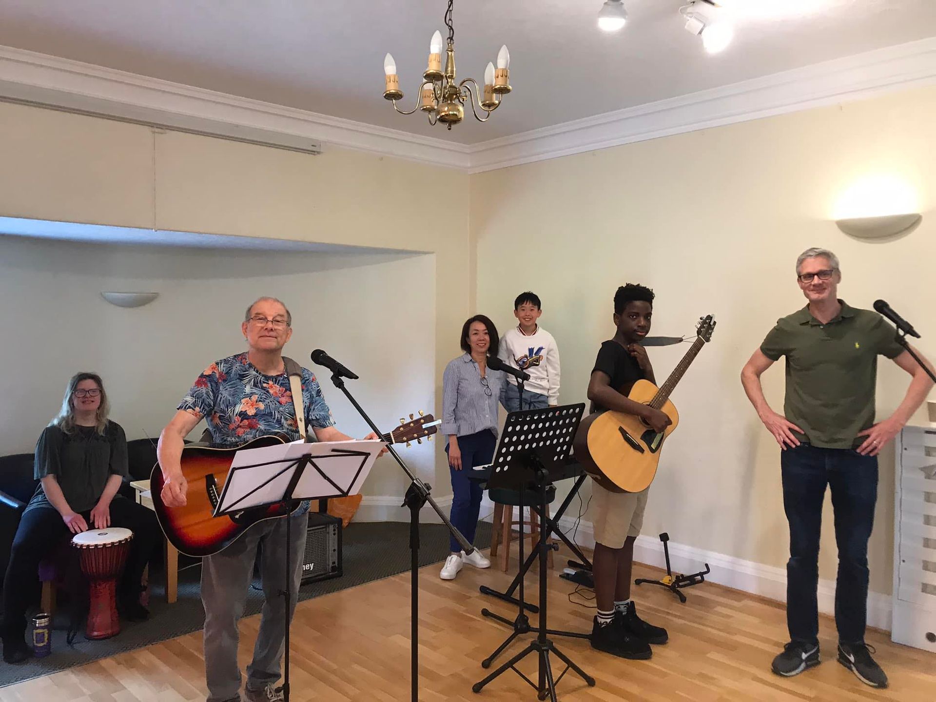A group of people practicing music in a studio; two play guitars, one drums, and others stand near music stands and mics.