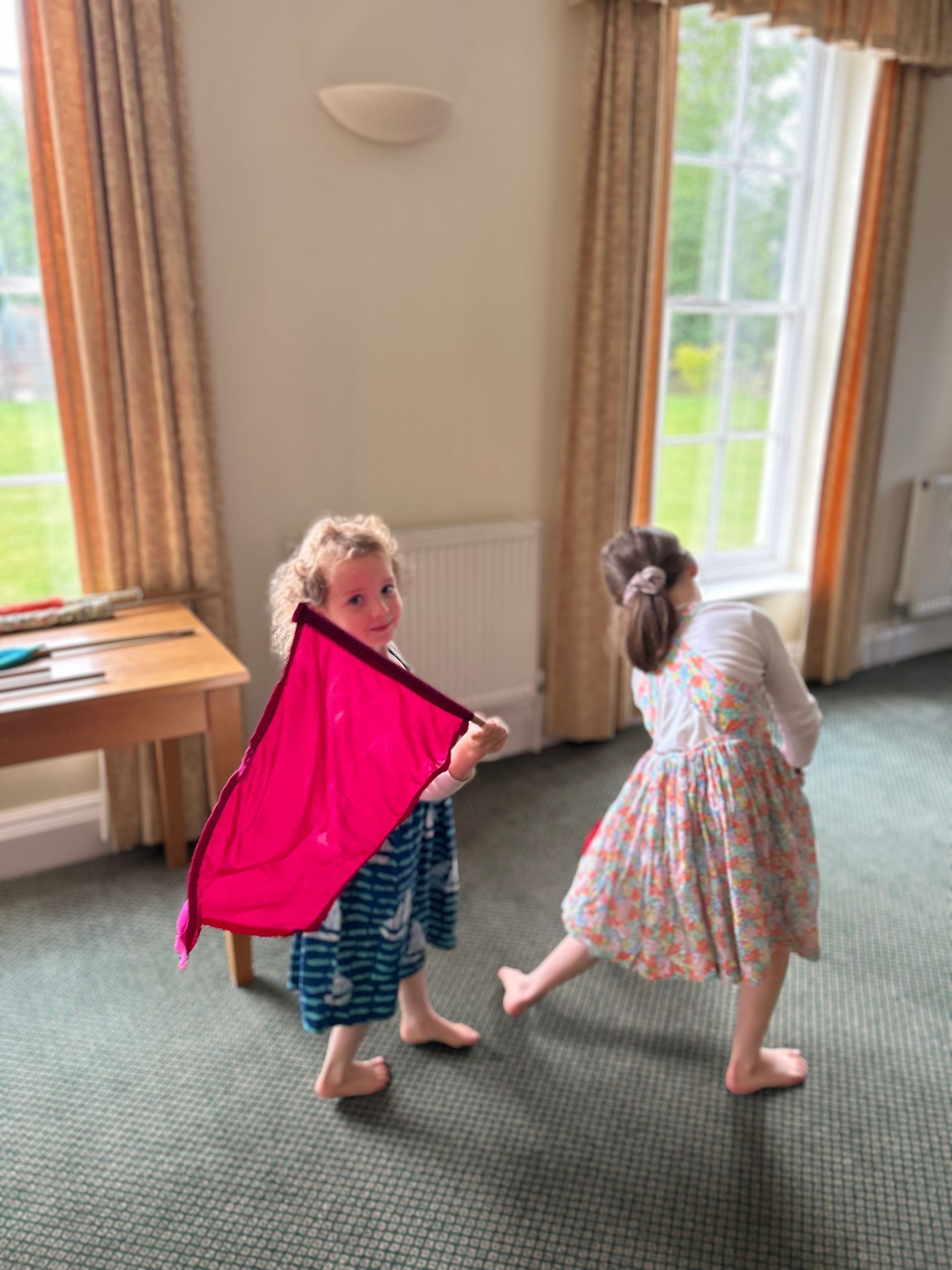 Two children play indoors near windows, one holding a bright pink cloth while the other stands in a patterned dress.