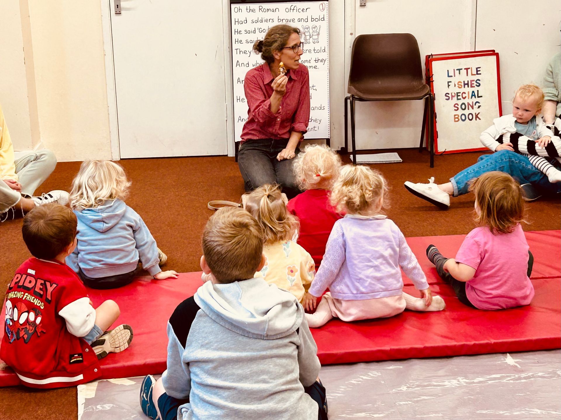 An instructor holds a small object while a group of children sits on a red mat in a room during a class activity.