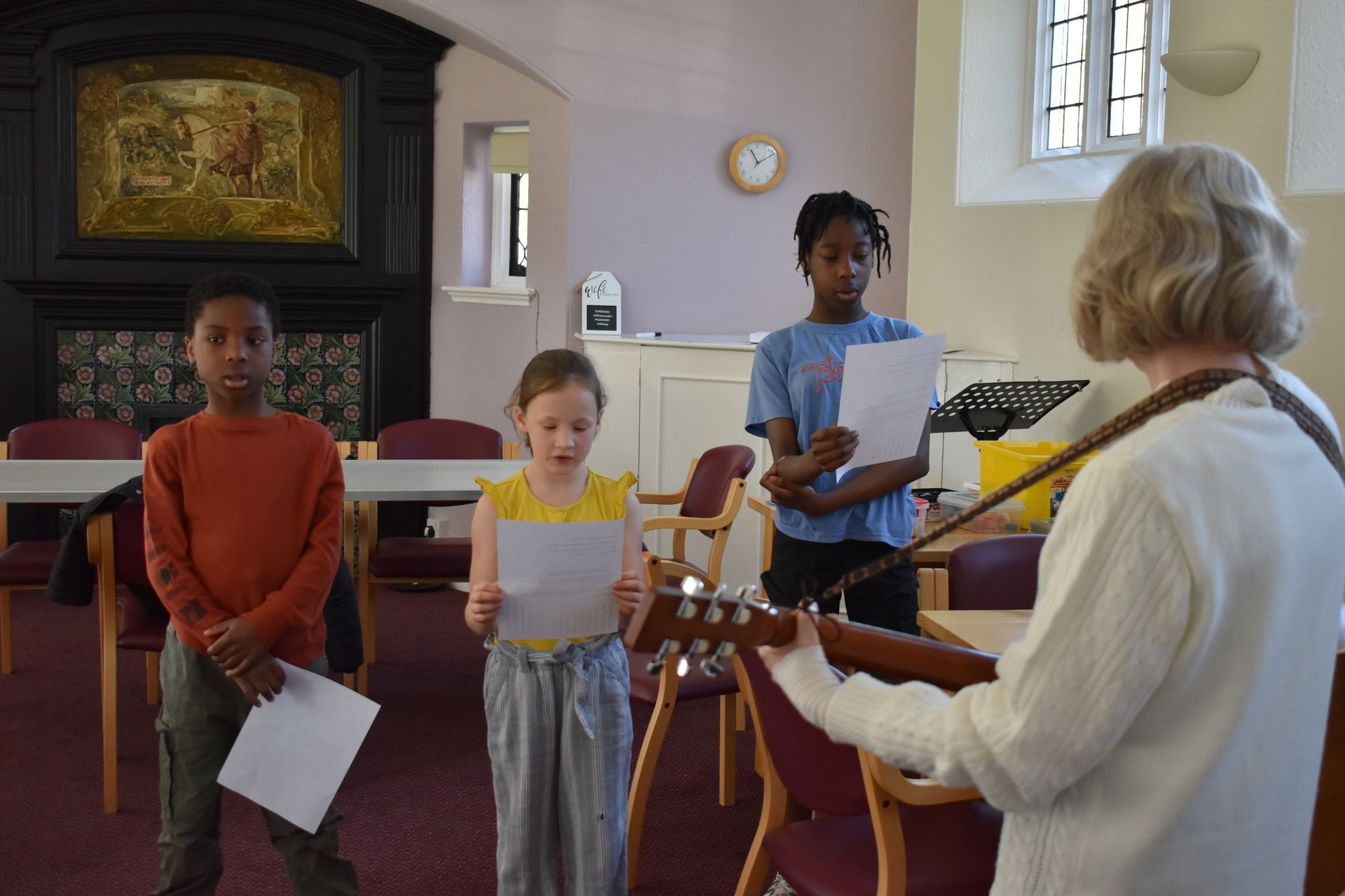 A group of children holding sheets of paper sing with an adult playing an acoustic guitar in a community hall.