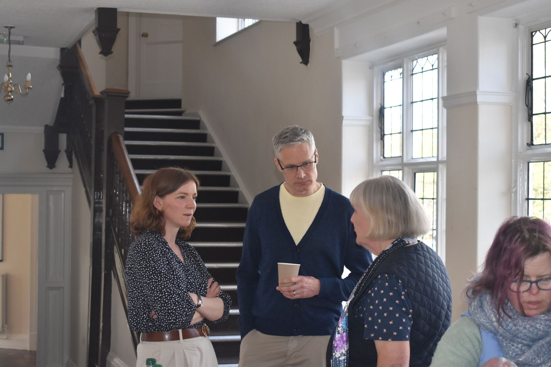 Four people stand in a bright, high-ceilinged room with a staircase, conversing and holding a drink.