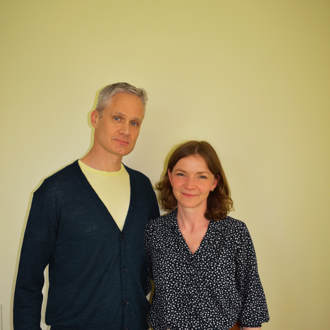 A man in a dark cardigan and yellow shirt stands next to a woman in a patterned dark top against a plain wall.