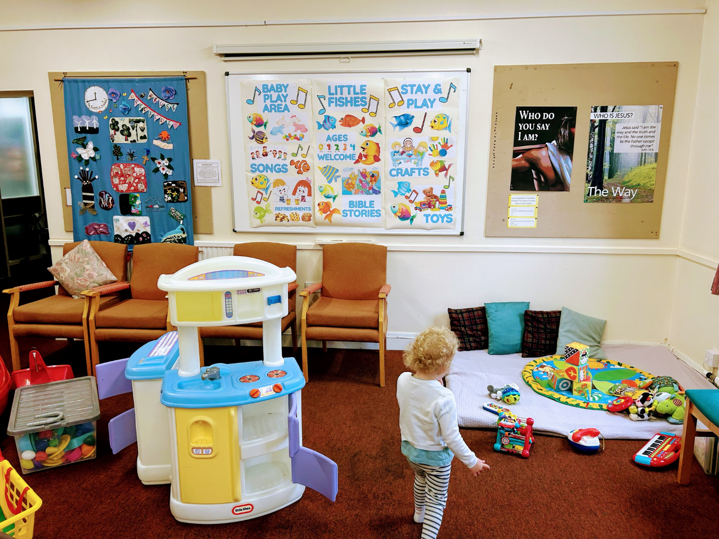 A toddler stands in a carpeted play area with a toy kitchen, chairs, a mat with toys, and posters on the walls.