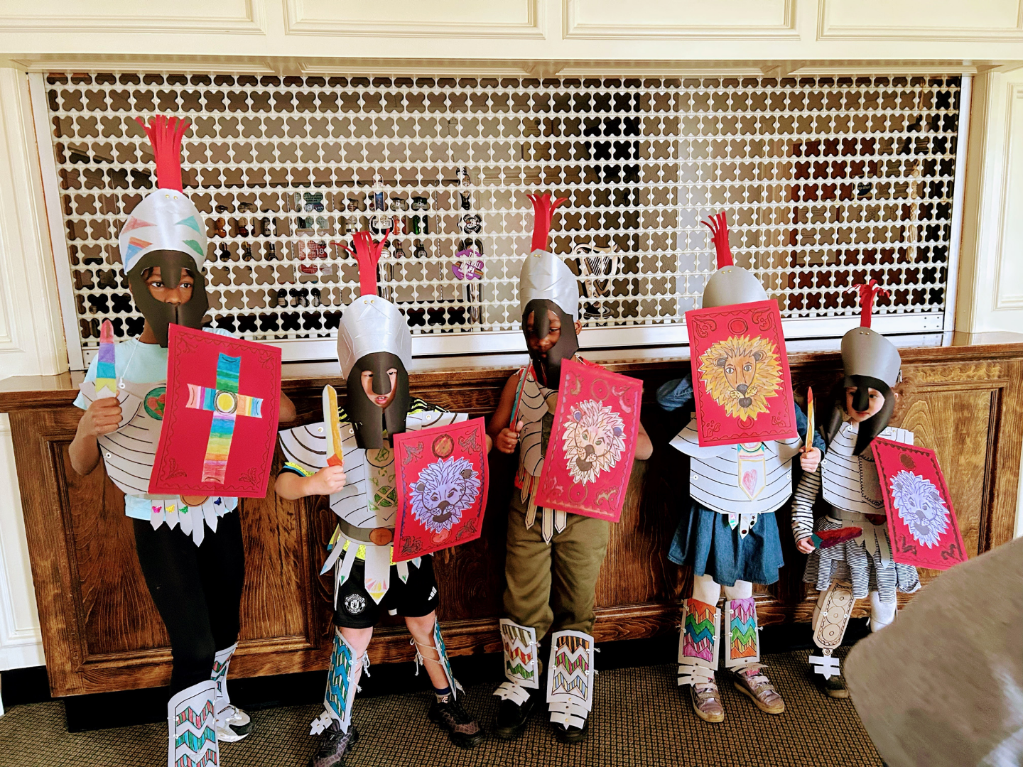 Five children in homemade knight armor and helmets stand in a line holding colorful shields inside a wood-paneled room.