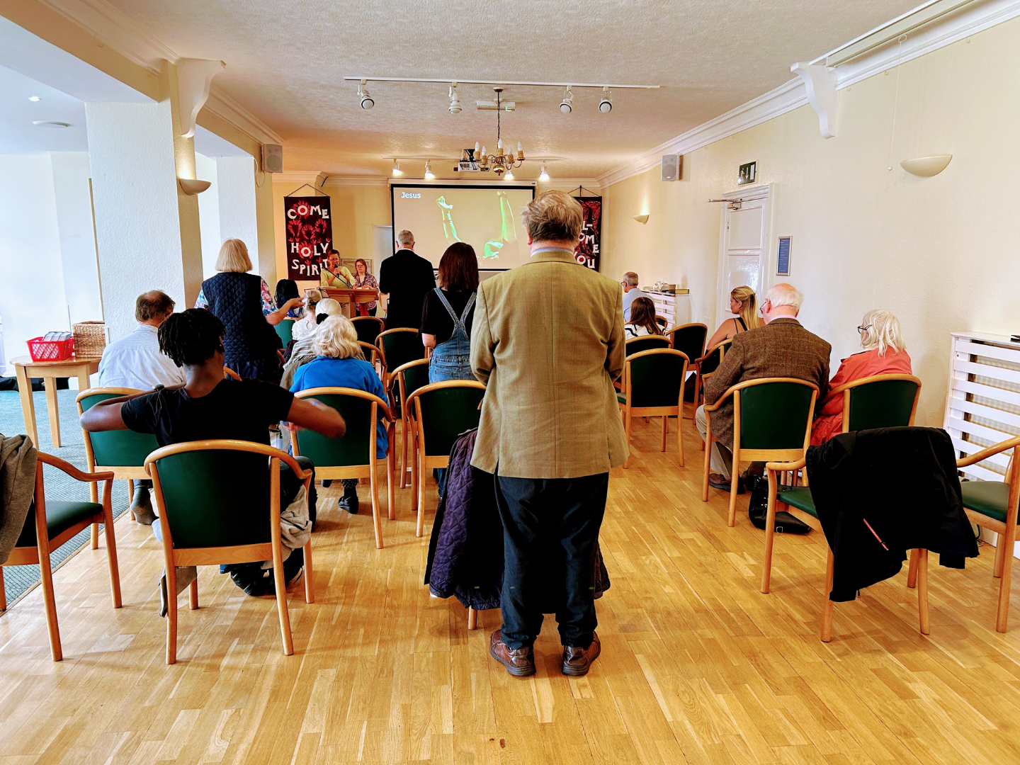 A group of people sit in chairs arranged in a room, facing a screen and a person standing in the foreground.
