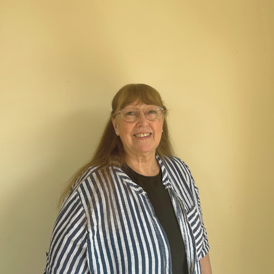 A smiling person wearing glasses and a blue and white striped shirt stands against a plain, light-colored background.