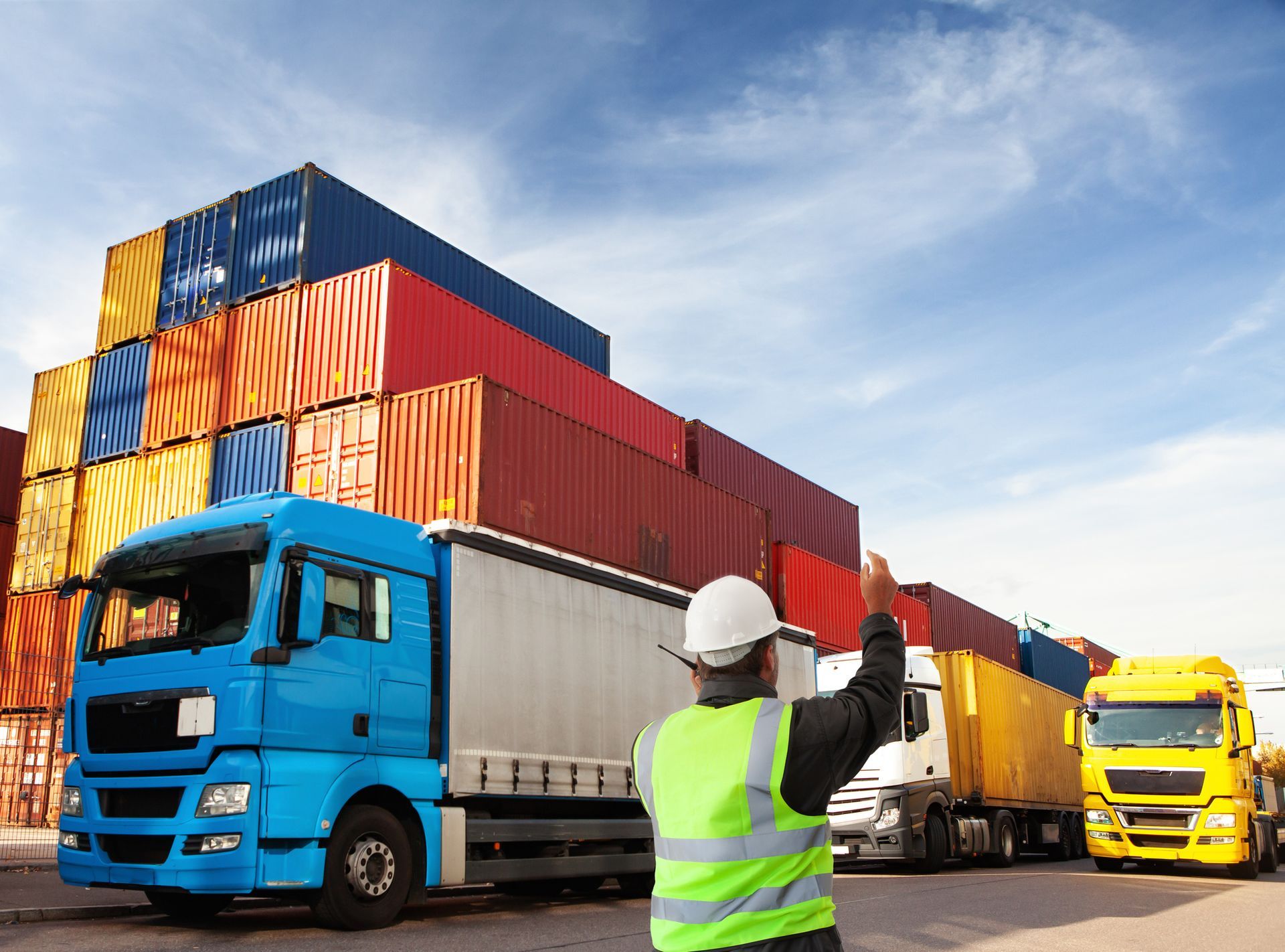 Worker directs trucks in a shipping yard filled with colorful cargo containers.
