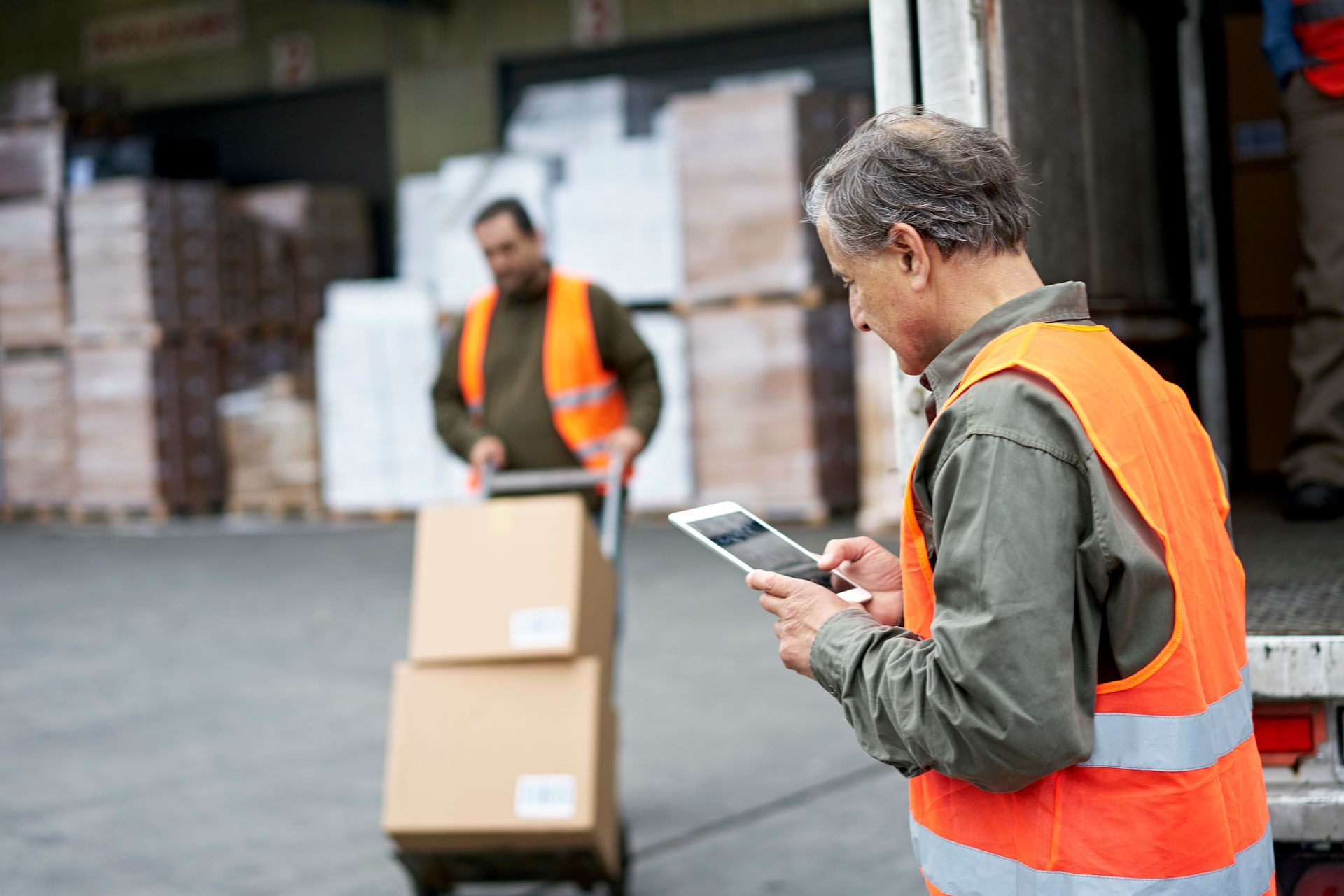 Man in orange vest uses a tablet near a loading dock. Another man loads boxes onto a truck. Man in orange vest uses a tablet near a loading dock. Another man loads boxes onto a truck.
