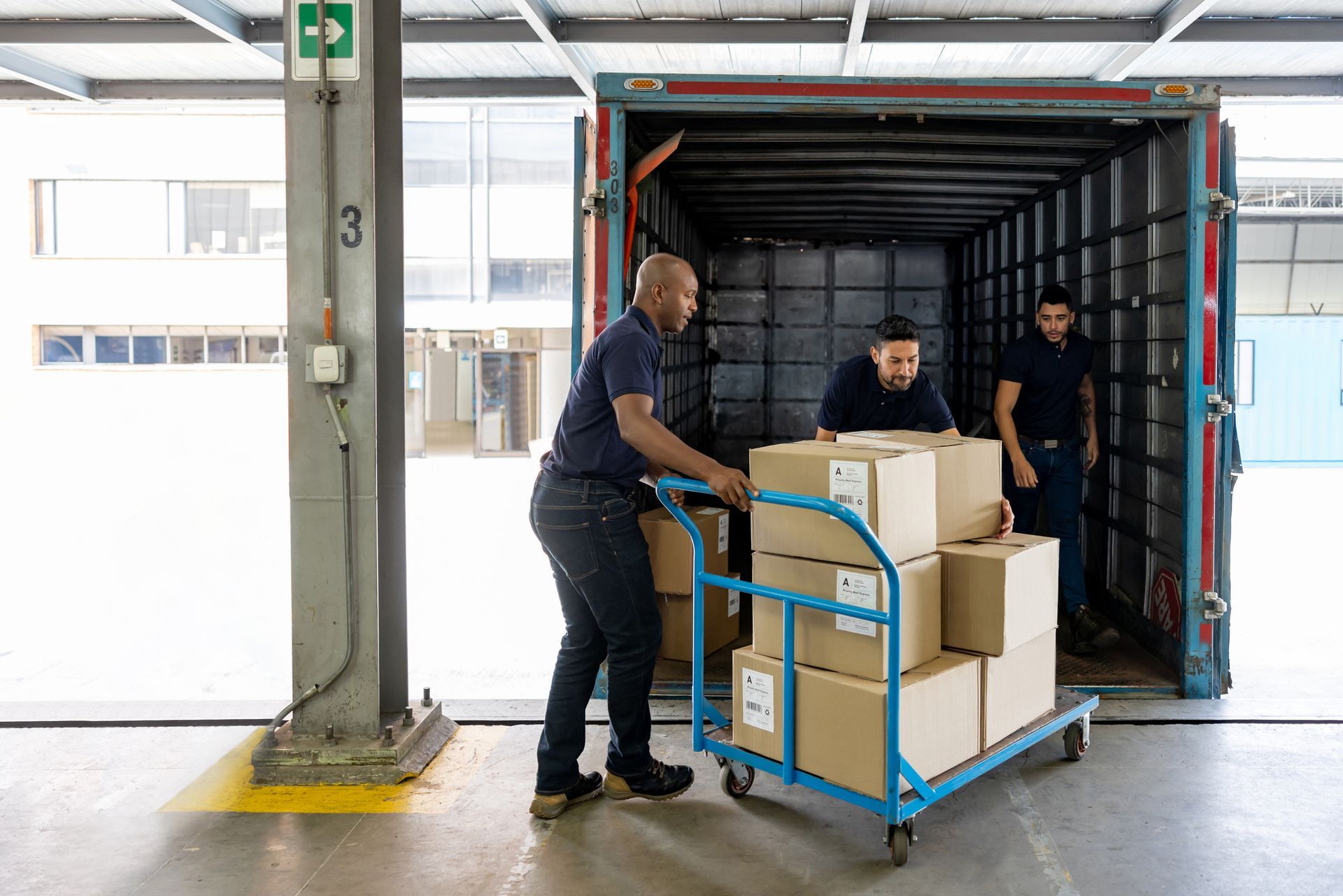 Three workers loading cardboard boxes from a cart into a shipping container at a warehouse. Three workers loading cardboard boxes from a cart into a shipping container at a warehouse.