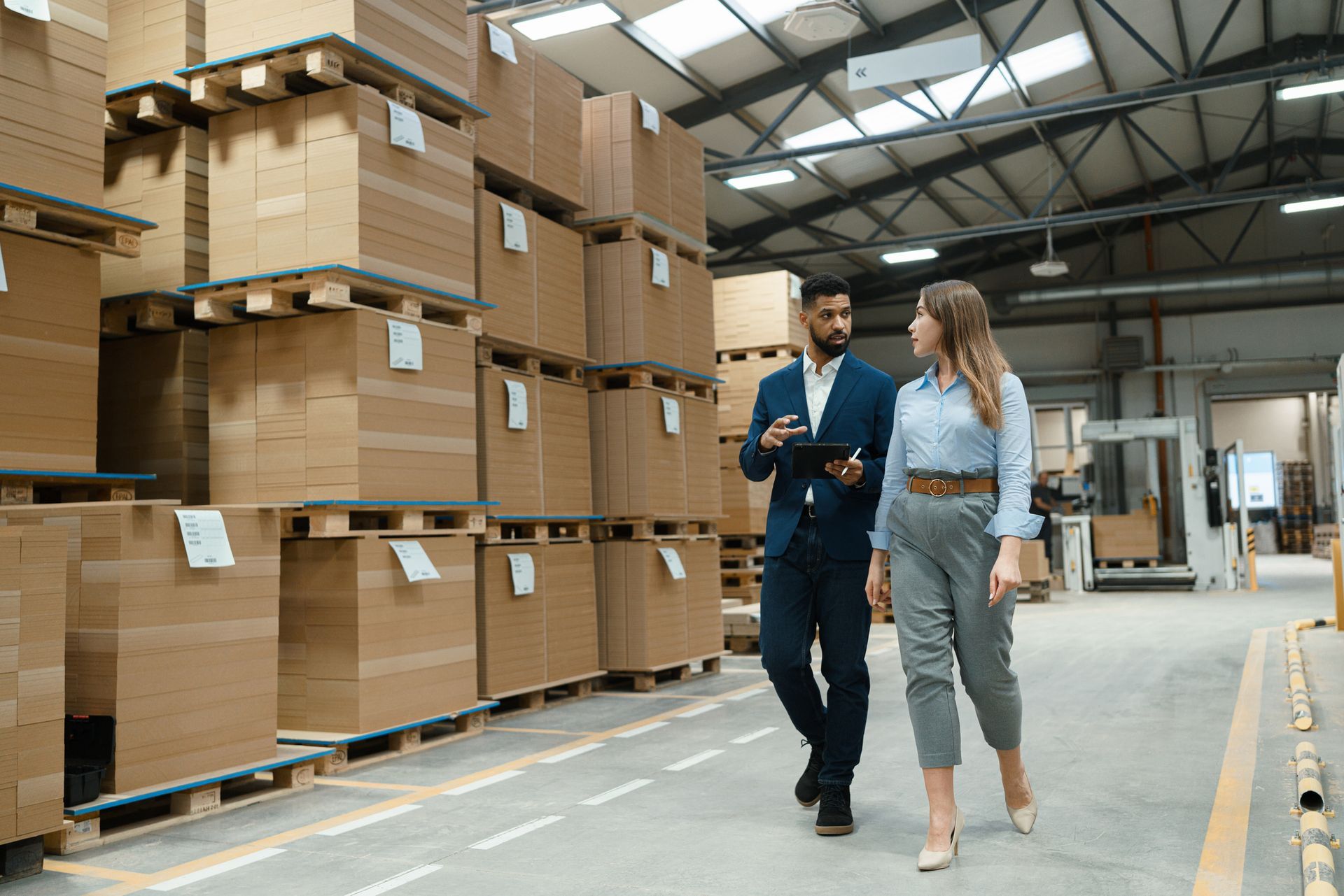 Man and woman walking and talking in a warehouse, surrounded by stacked boxes on pallets. Man and woman walking and talking in a warehouse, surrounded by stacked boxes on pallets.