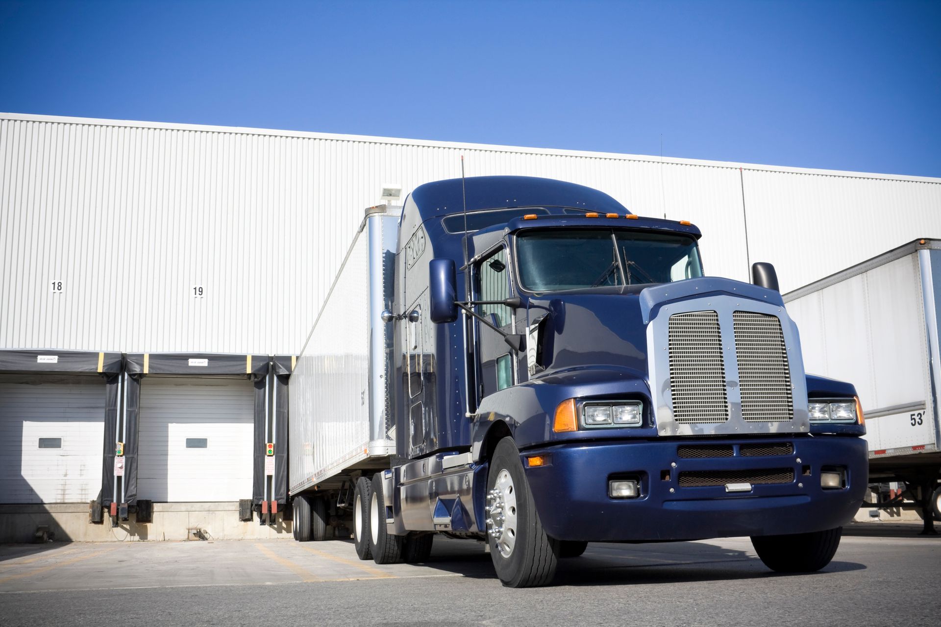 Dark blue semi-truck at a loading dock, white trailer, blue sky, and warehouse in background.