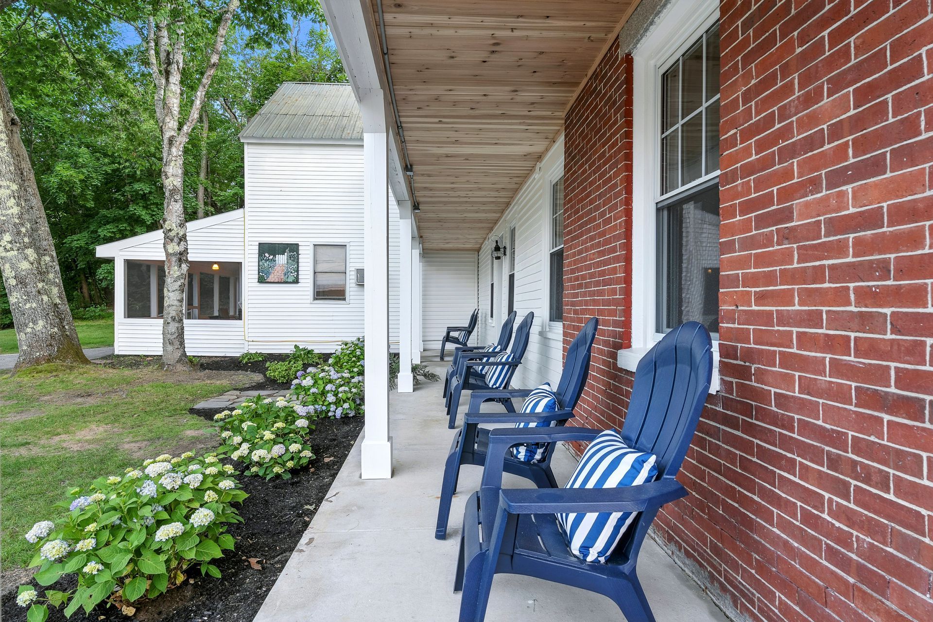 Porch of a brick building with blue chairs. White trim, wood ceiling, green plants, and trees.