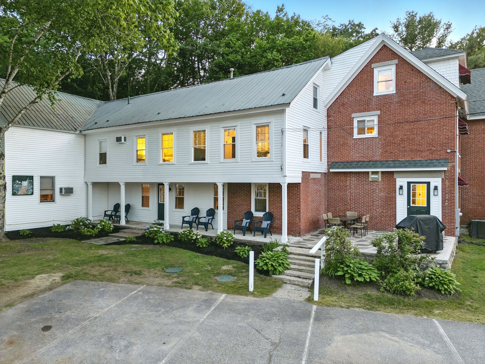 Two-story white and red brick building with metal roof; porch with chairs, green lawn, trees.