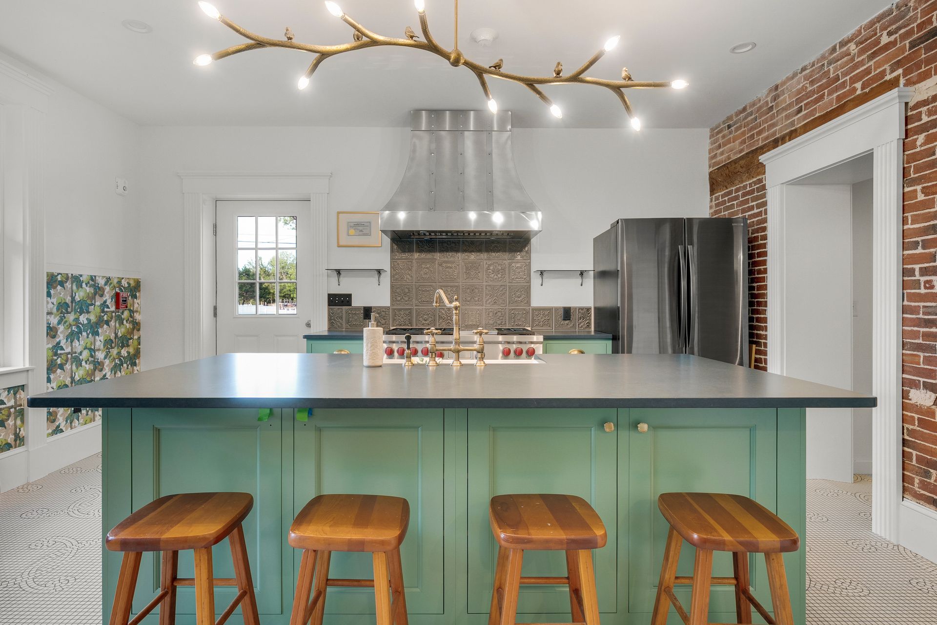 Green kitchen island with wooden stools, stainless steel appliances, brick wall, and decorative chandelier.
