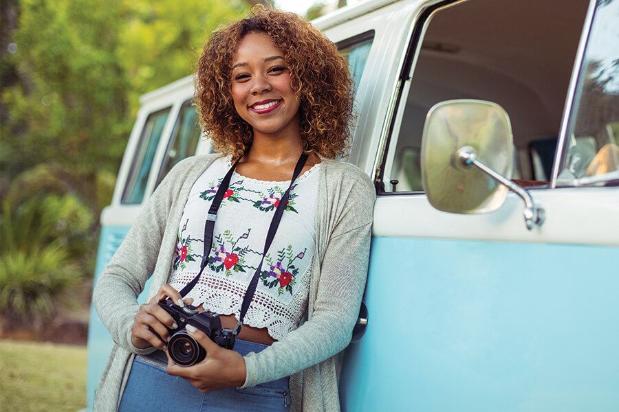 Smiling person holding a camera beside a light blue van outdoors