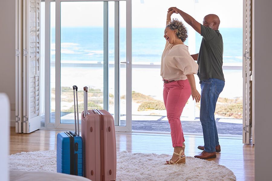 Couple by oceanfront doors, one adjusting the other's hair near two suitcases