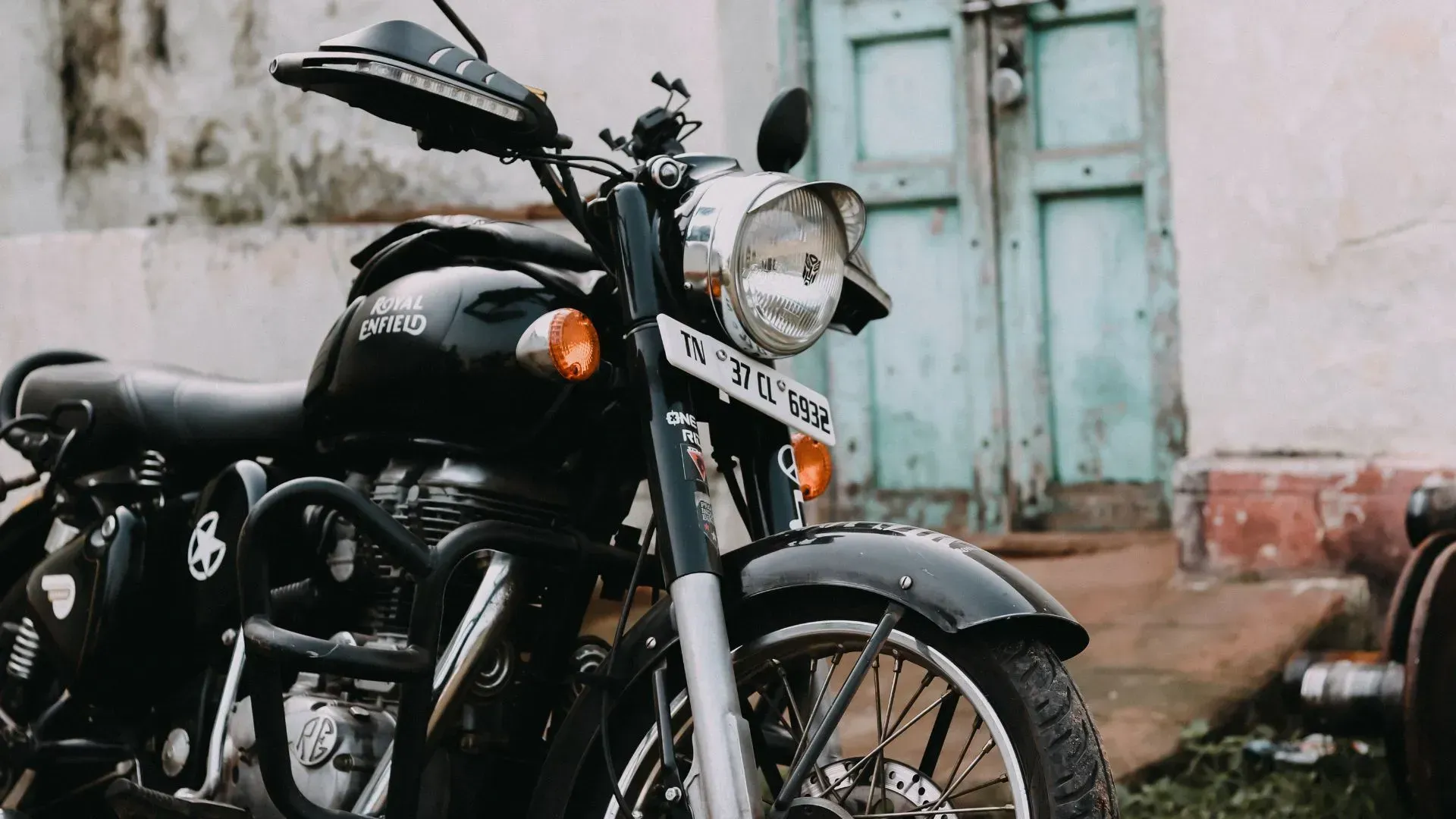 Motocicleta preta estacionada em frente a um prédio desgastado com porta azul-esverdeada e parede branca.