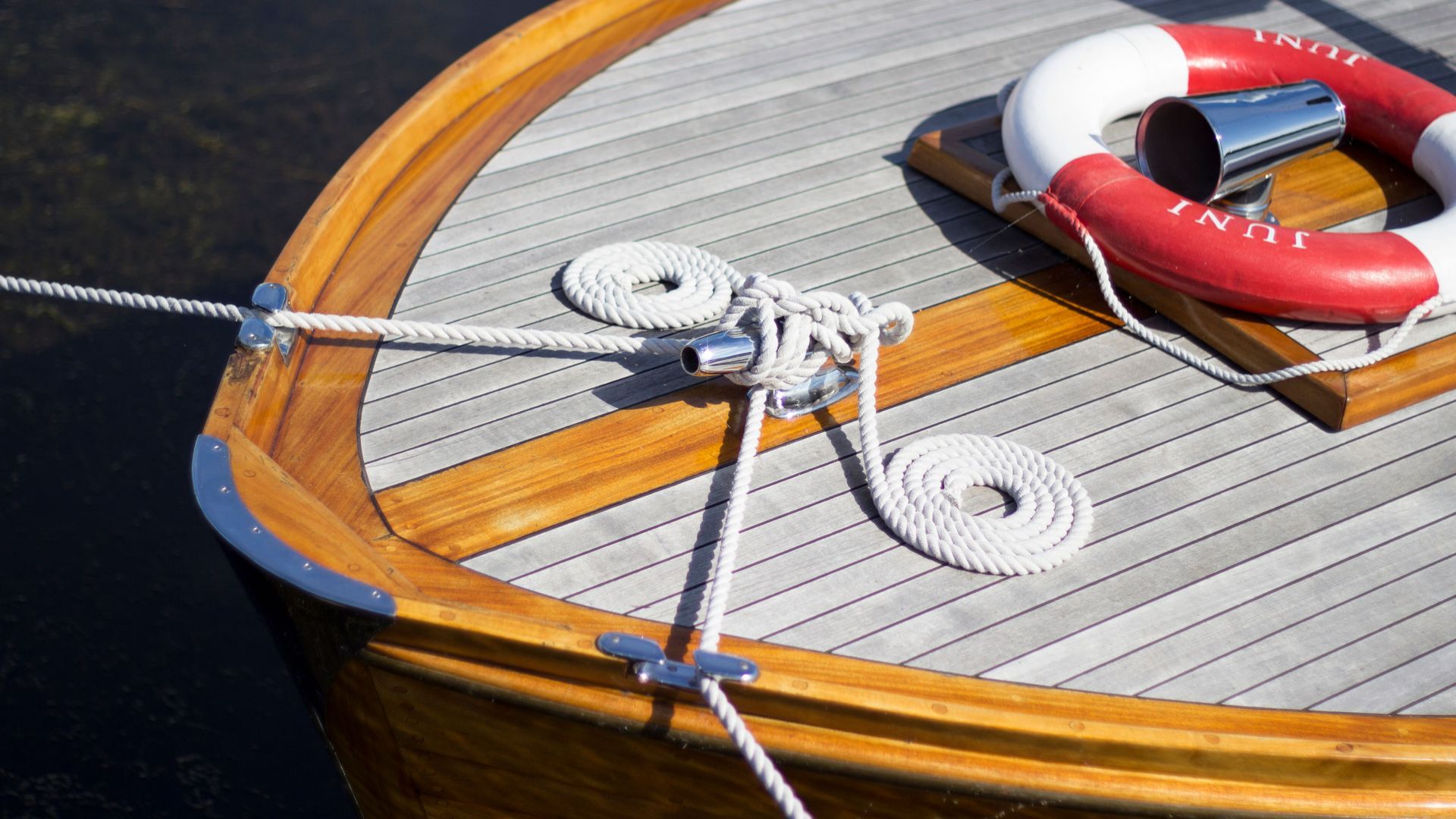 A wooden boat deck with ropes, white cleats, and a red-and-white life ring near the railing.
