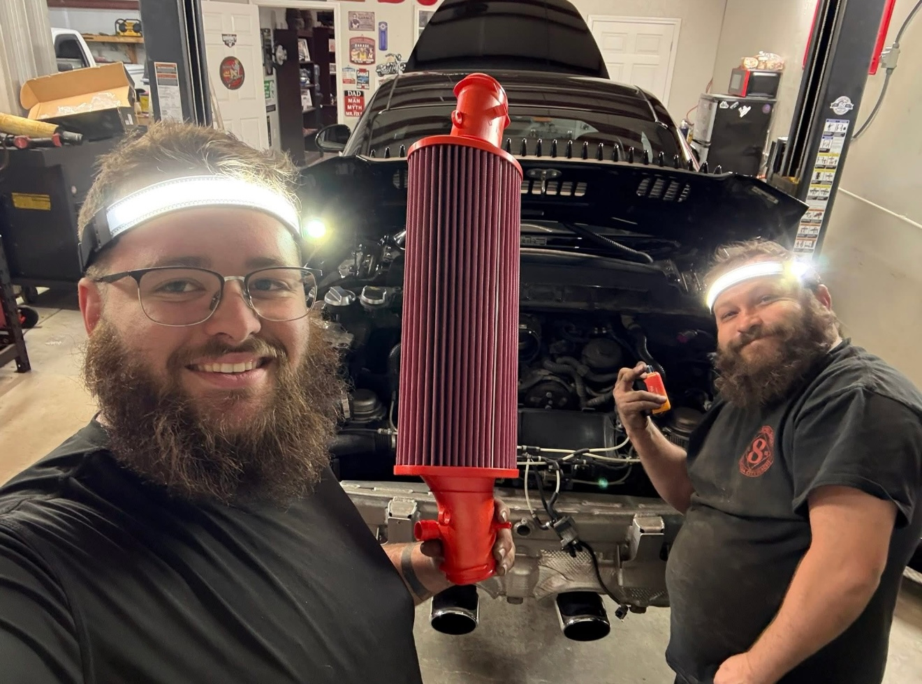 Two people wearing headlamps smile in a workshop, holding a large, red, cylindrical automotive air filter.  | Bucks Auto Garage & Tire