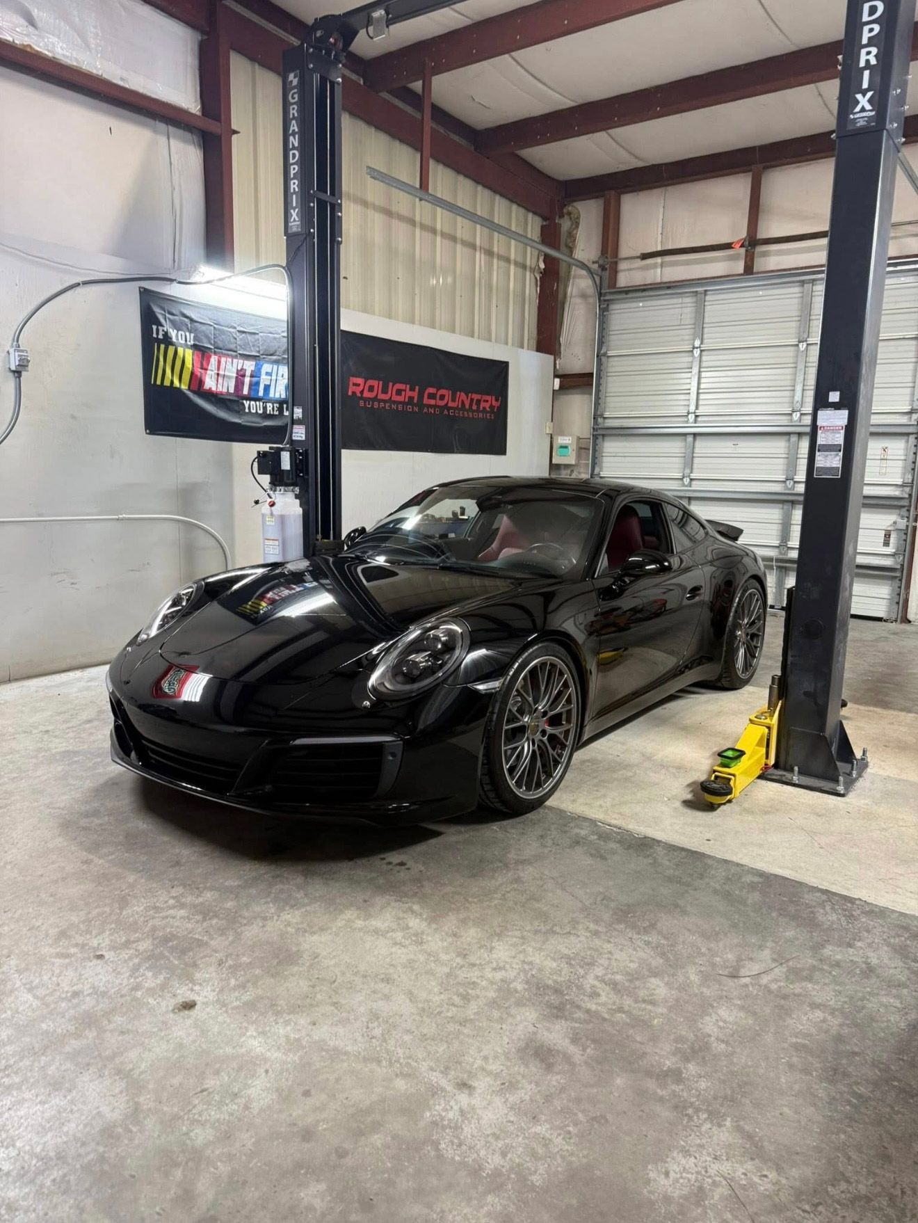 A black Porsche sports car is parked inside an automotive garage between the vertical support beams of a two-post car lift. | Bucks Auto Garage & Tire