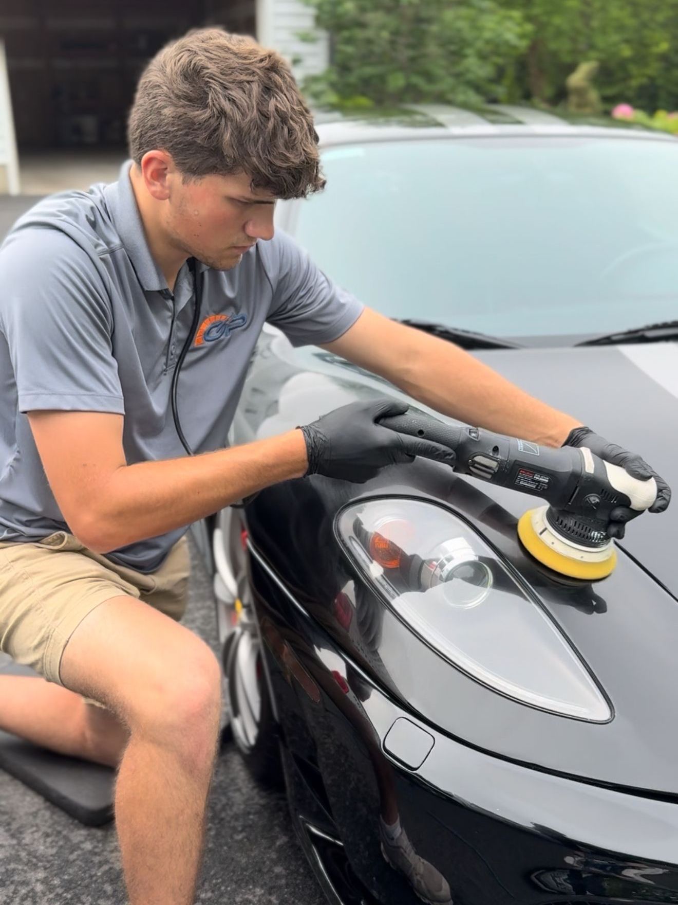 Person polishing a black car hood with a yellow buffer outdoors.
