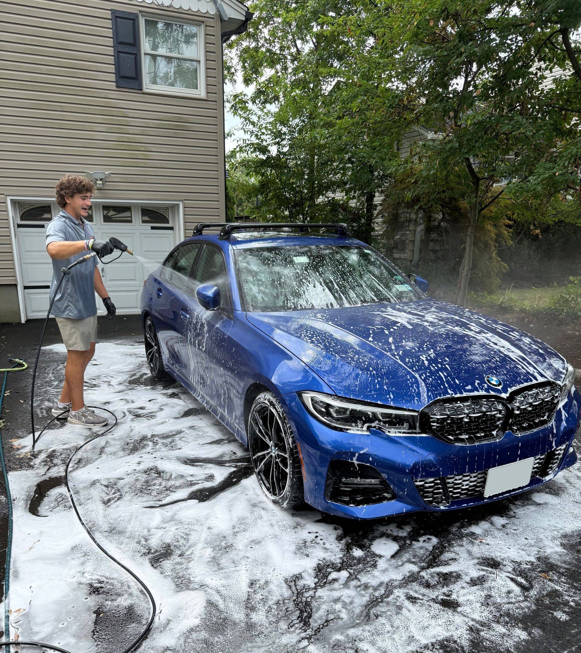 Person washing a blue BMW sedan with foam in a driveway beside a house