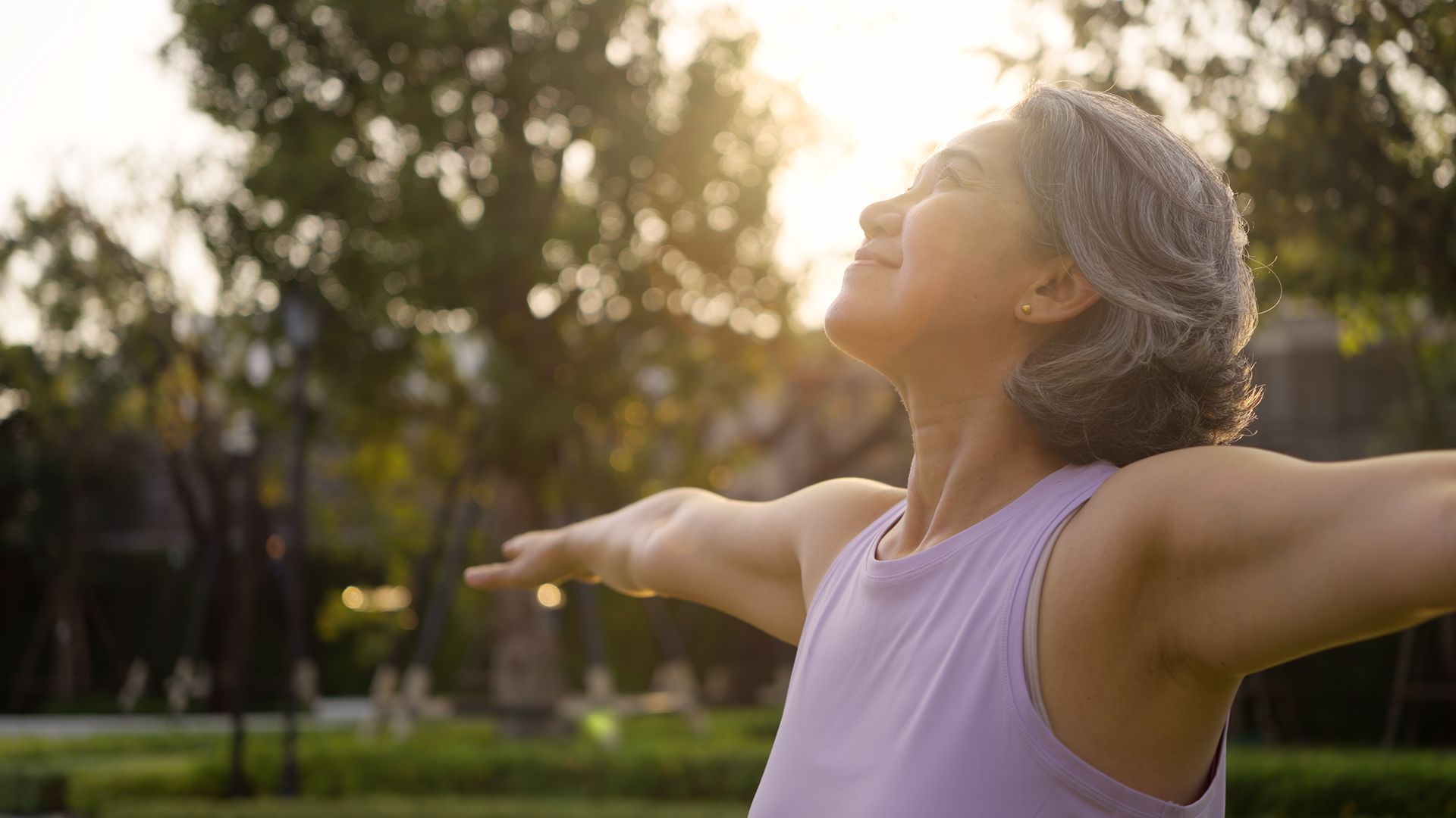Person in a lavender top with arms outstretched, enjoying sunlight in a park.