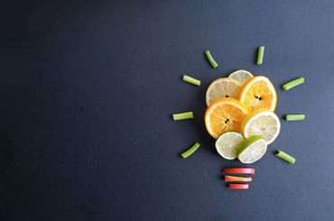Citrus slices and cucumber arranged as a lightbulb on a dark background, with red staples at the base