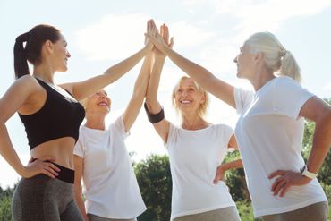 Four women in athletic wear high-five outdoors under a bright blue sky.