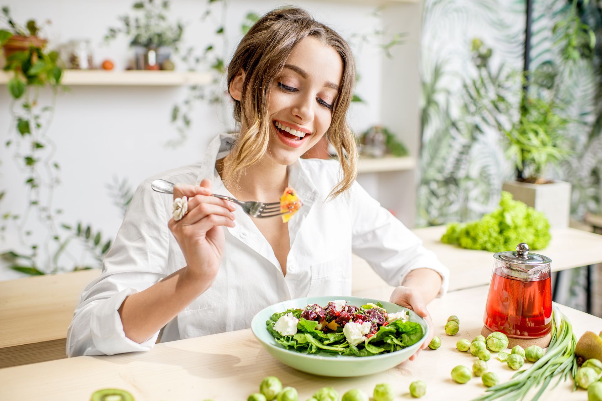 Woman smiles, eating salad in kitchen. Green salad in bowl, fruit on table. Wooden table, leafy background.
