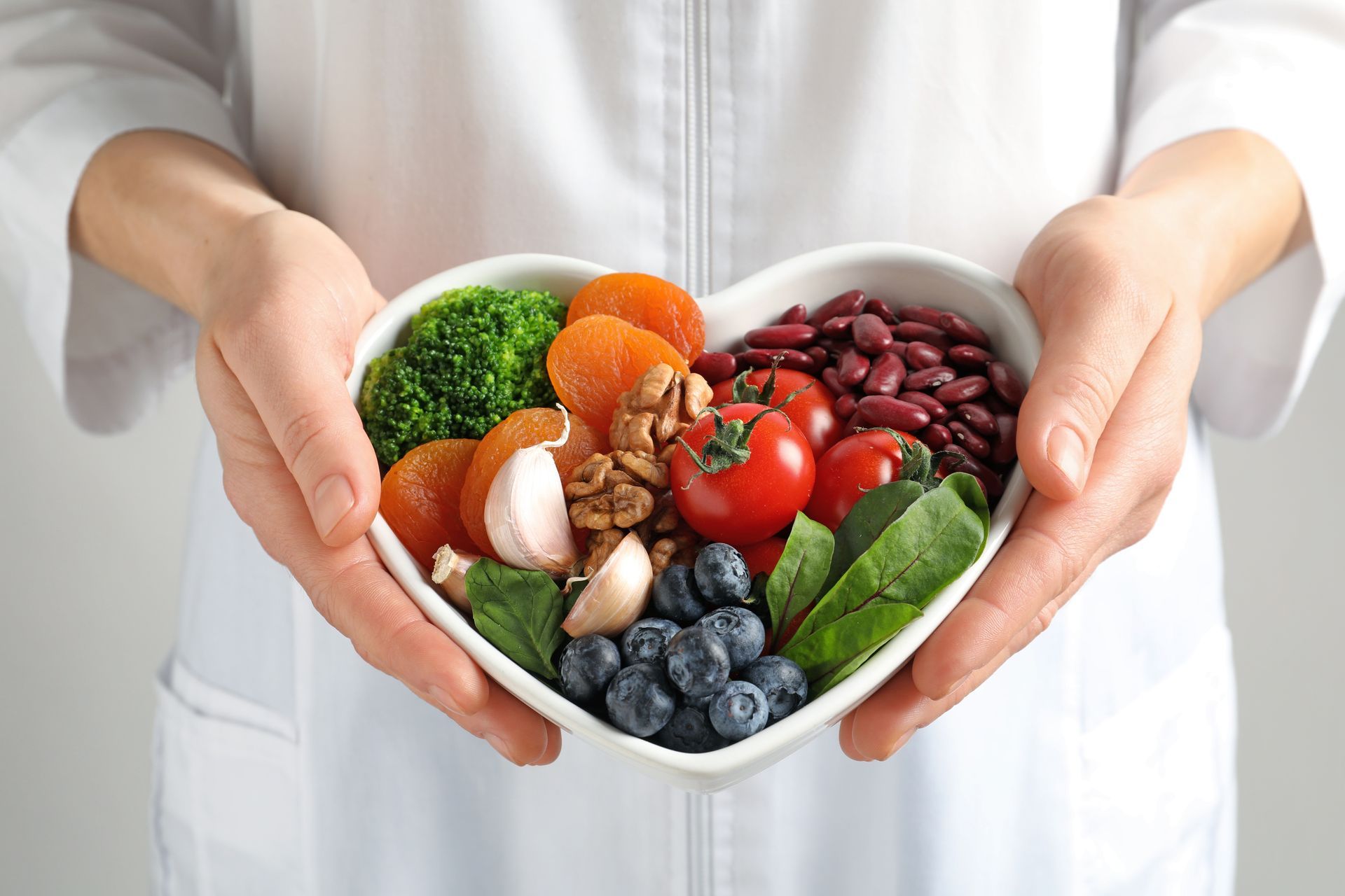 Hands holding a heart-shaped bowl filled with colorful, healthy foods like vegetables, beans, and berries.