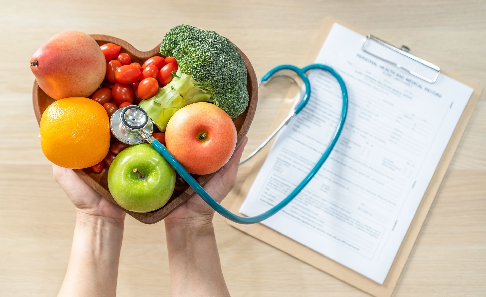 Hands holding heart-shaped bowl of fruit and vegetables; stethoscope and clipboard on a wooden surface.