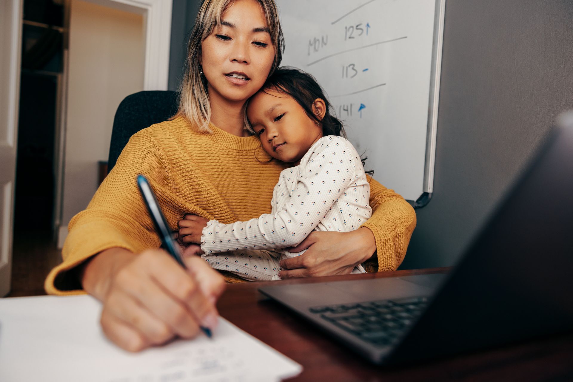Woman writing with a pen, holding a child who is resting their head on her shoulder. Laptop and whiteboard are nearby.