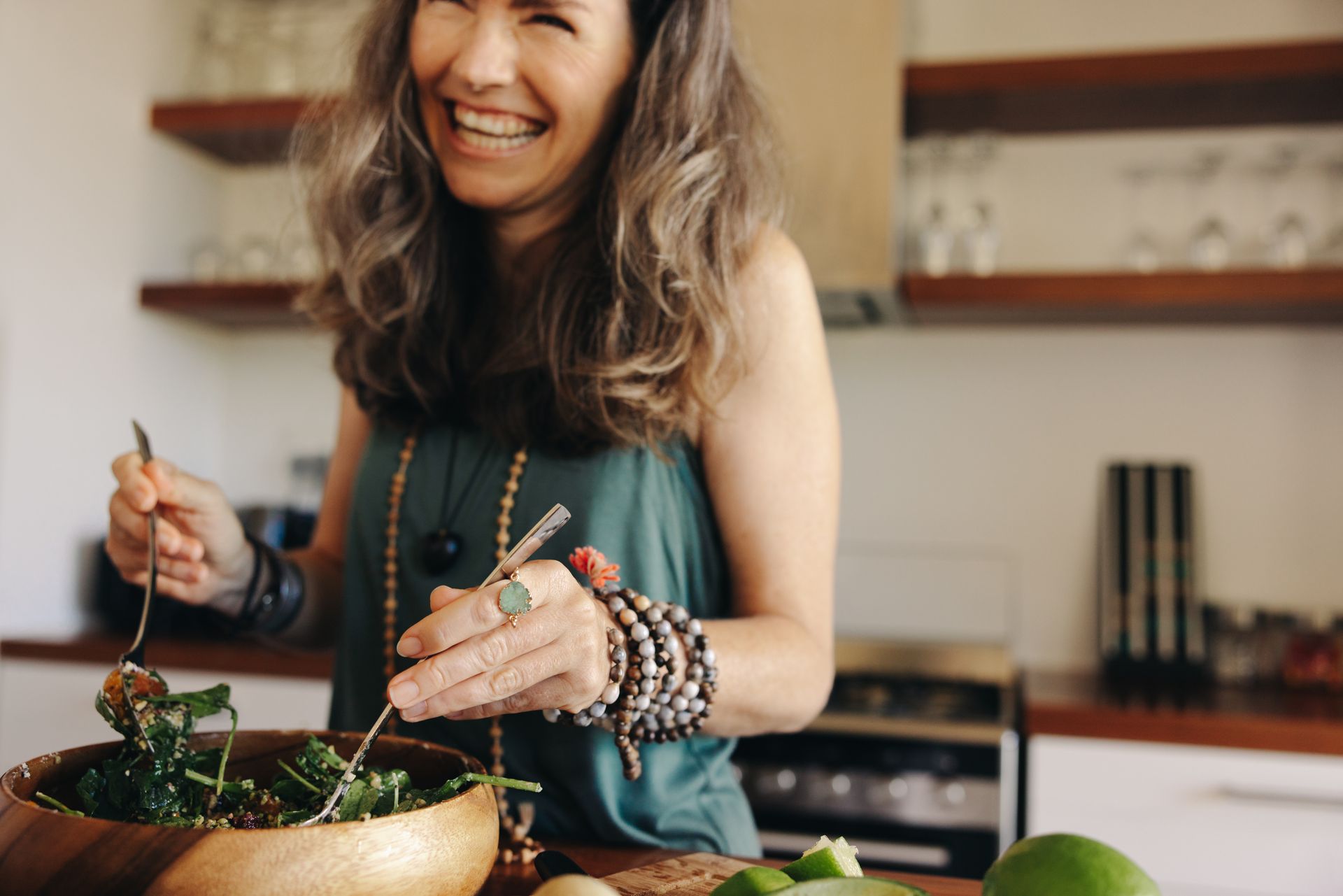 Woman in kitchen smiles, tossing salad with a spoon and fork, wooden bowl, bracelets.