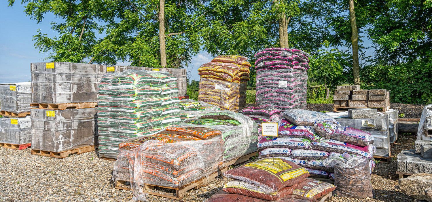 Stacks of bundled materials and sacks piled outdoors in a yard, with trees and stone walls behind.
