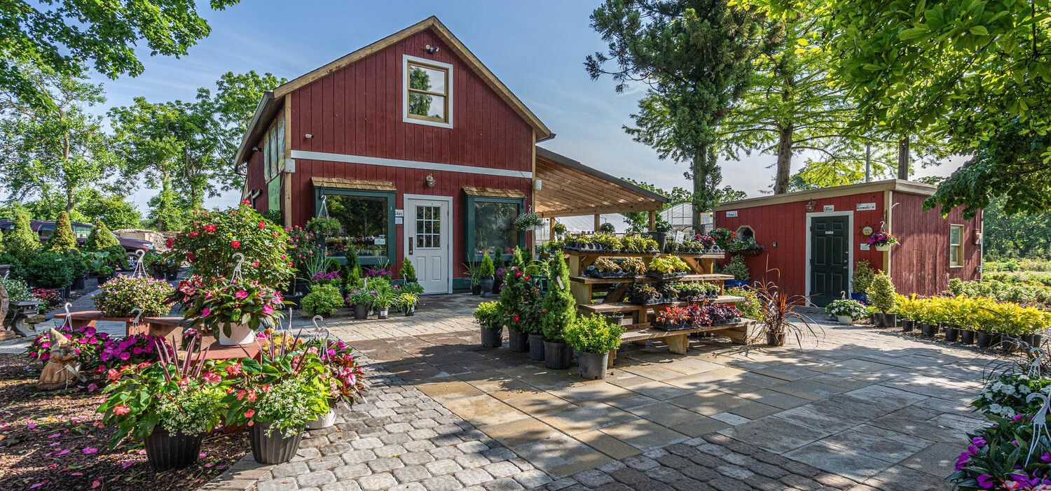 Red farmhouse with white trim, flower gardens, and a stone patio on a sunny day