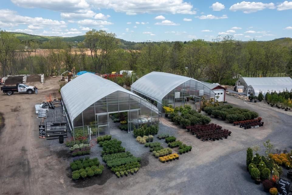 Aerial view of a greenhouse nursery with rows of potted plants and trees outdoors under a partly cloudy sky