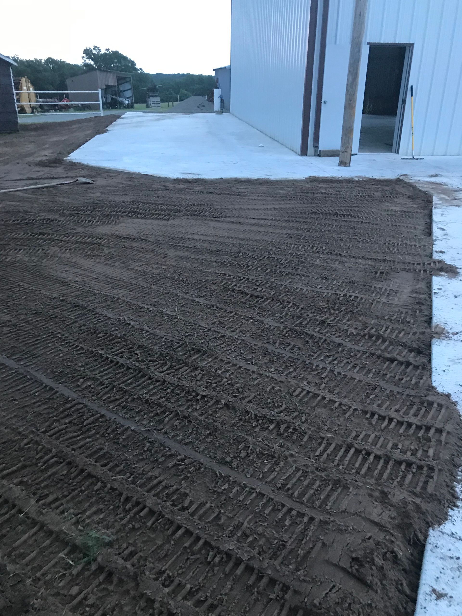 A large area of dark, tilled soil with tire tracks next to a white metal building and a concrete apron.