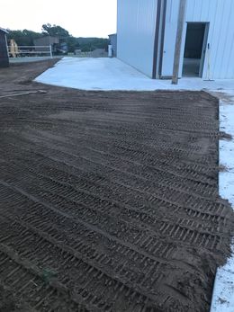 A dirt construction area with heavy equipment tire tracks in front of a white metal building with a concrete apron.