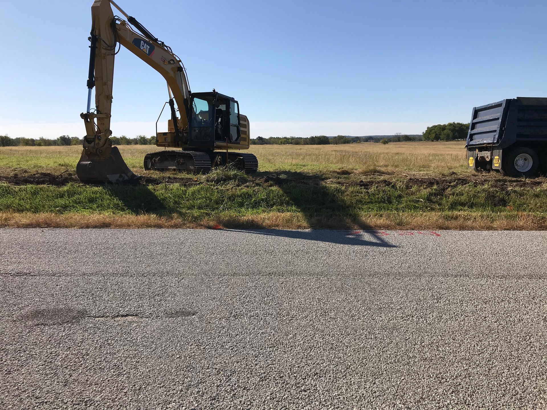 A yellow Caterpillar excavator digging in a grassy field next to a dump truck and a gravel road under a clear blue sky.