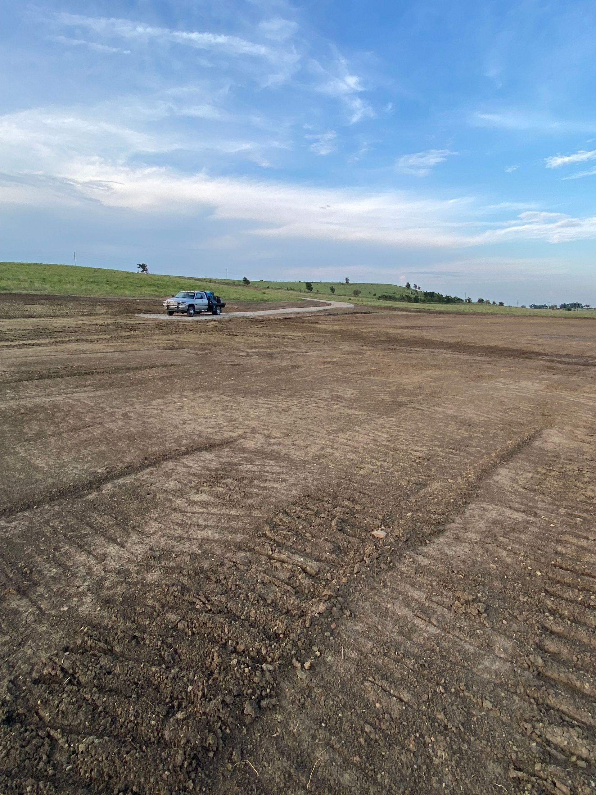 A blue vehicle drives across a large, dirt-covered construction field under a bright, cloudy sky.