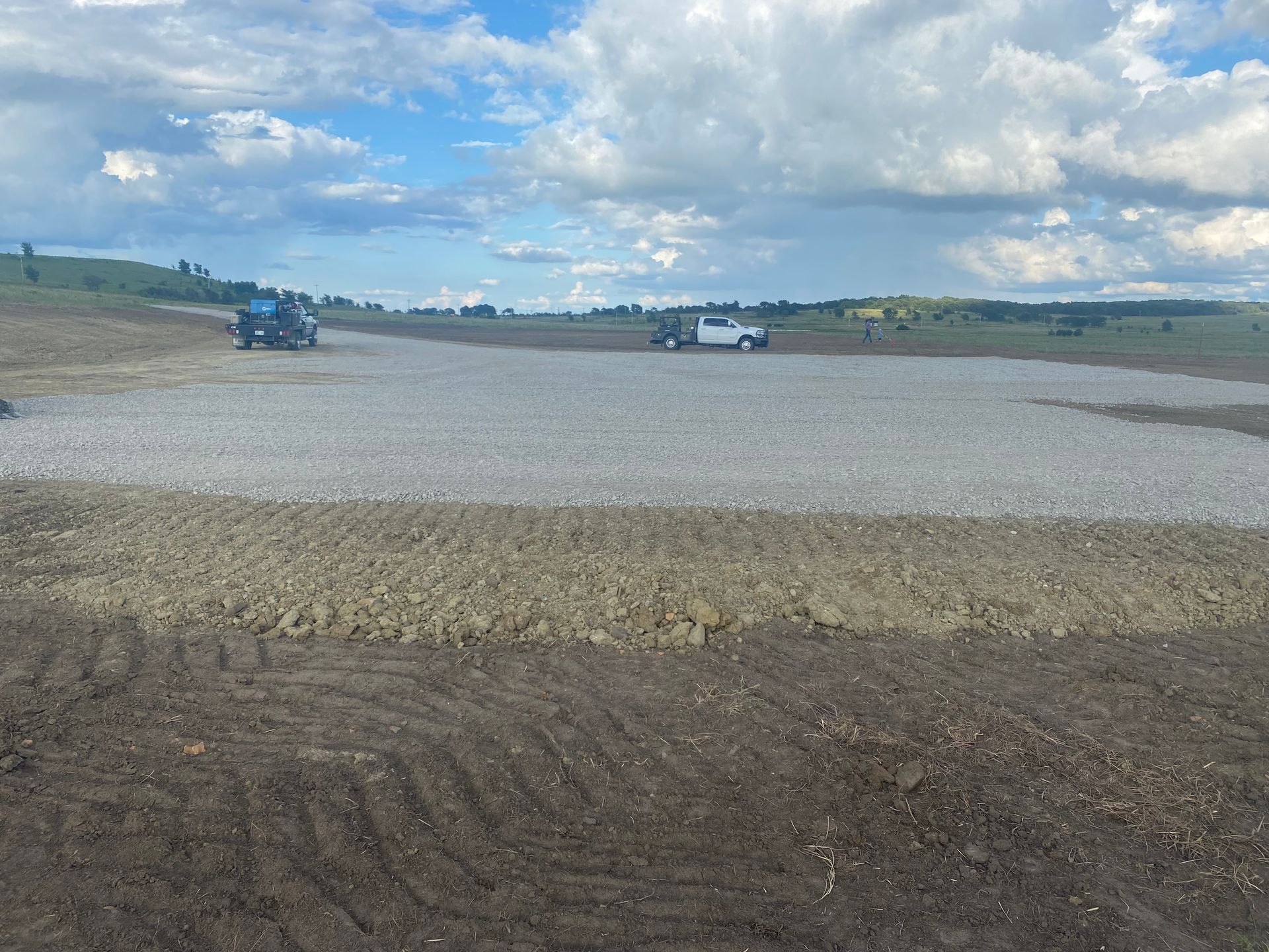 A gravel pad under construction in a large field with two pickup trucks parked in the distance under a cloudy sky.
