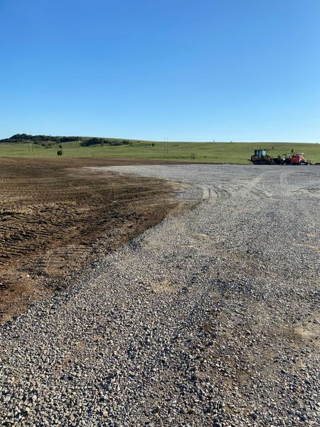 A gravel road stretches across a rural landscape under a clear blue sky, with heavy machinery visible in the distance.