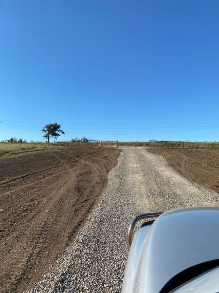 A view from a vehicle driving down a gravel road through a cleared, grassy field under a clear blue sky.