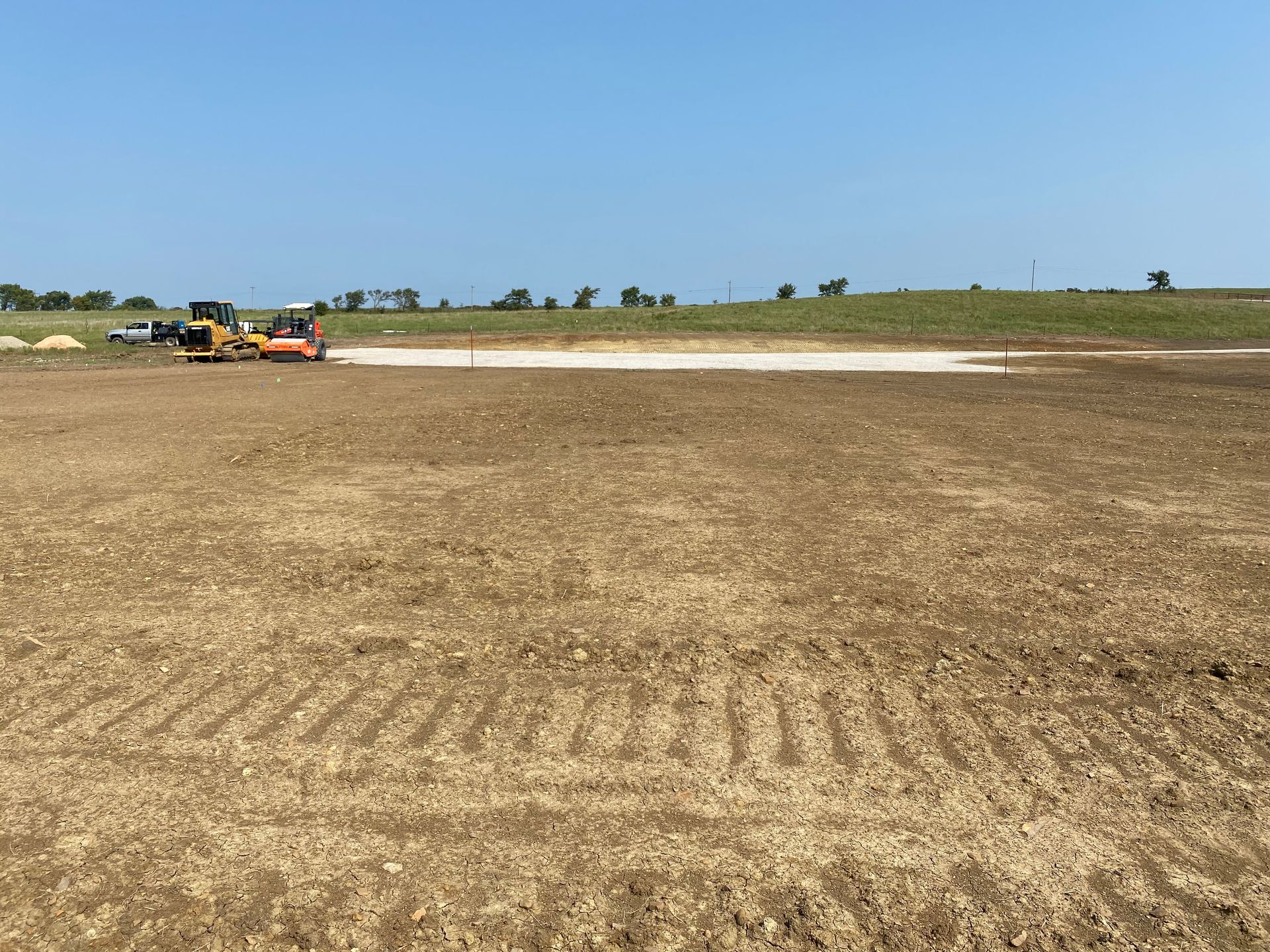 Construction equipment levels a patch of light-colored gravel on a large, flat, excavated dirt field under a blue sky.
