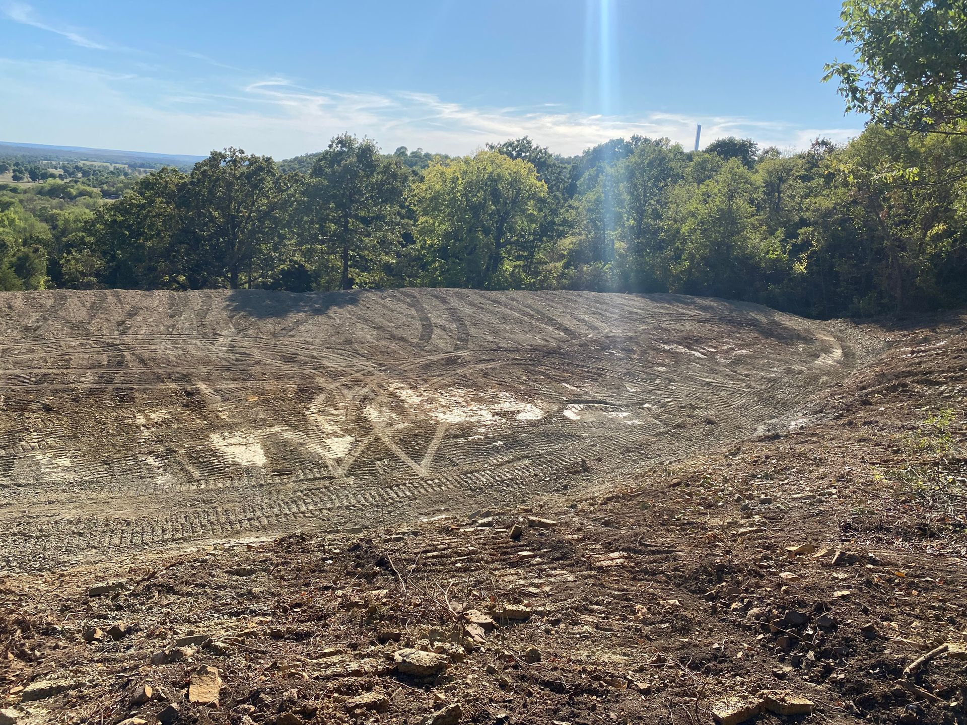A cleared, uneven dirt lot with light-colored soil and dark mulch, bordering a line of trees under a bright blue sky.