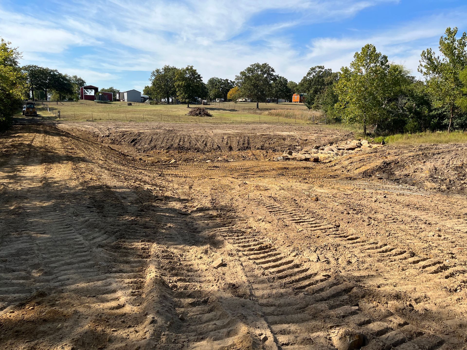 A cleared, dirt-filled plot of land with tire tracks in the foreground, facing a field with trees under a blue sky.