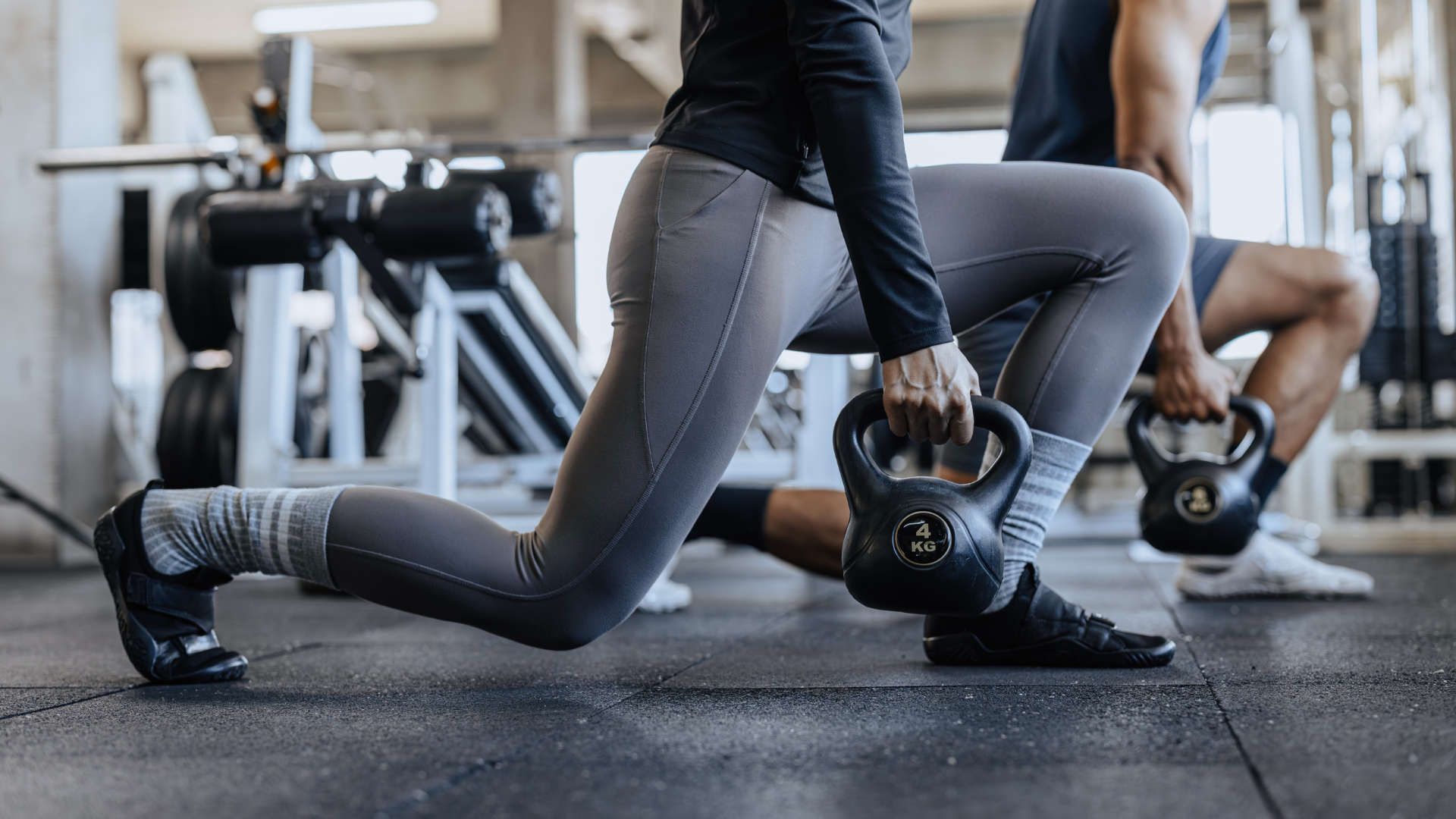 People lunging with kettlebells in a gym, wearing athletic gear on a rubber floor.